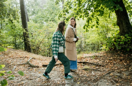 Twee vrouwen wandelen in een bos tijdens de herfst, omringd door groene en gevallen bladeren. Ze lachen en lijken een gesprek te voeren. Eén draagt een beige jas en een lange spijkerbroek, de ander een groen-witte geruite jas met groene broek en sneakers. (vergroot foto)