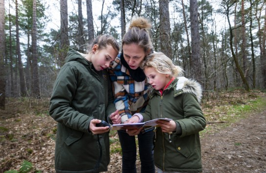 Drie personen in winterjassen bekijken samen een kaart en een GPS-apparaat in een bos. (vergroot foto)