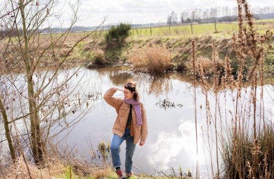 Vrouw in jas en sjaal kijkt over een riviertje in een open landschap met droge planten en gras. (vergroot foto)