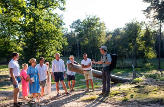 Groep mensen luistert naar gids in natuurgebied met bomen en zonlicht in de namiddag. (vergroot foto)