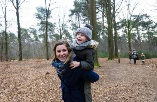 Vrouw draagt lachend kind op haar rug in bos met herfstbladeren, op de achtergrond lopen drie andere mensen. (vergroot foto)