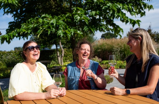 Drie vrouwen zitten buiten aan een houten tafel, lachen en drinken witte wijn in een zonnige tuin. (vergroot foto)