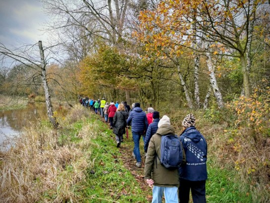 Groep mensen wandelt in herfstbos langs een smal pad naast een waterpartij onder bewolkte lucht. (vergroot foto)